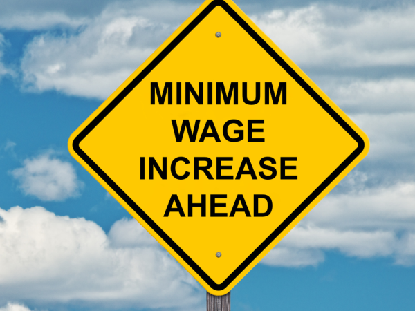 A blue sky with a scattering of white clouds and a yellow road sign with the words 'MINIMUM WAGE INCREASE AHEAD' in black text.