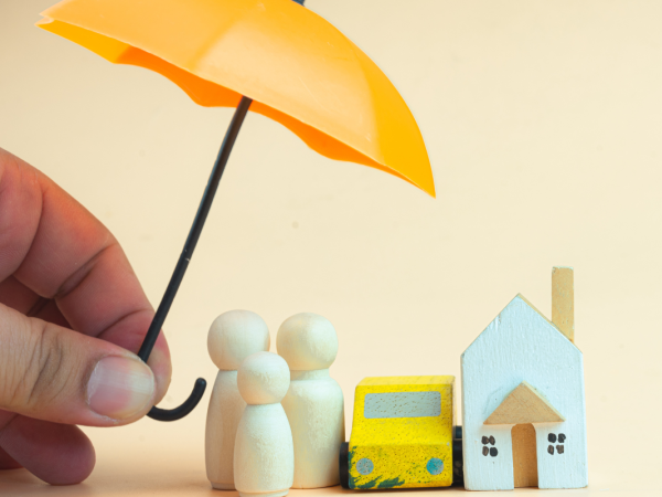 A small model home with a small family and car, a person is holding an umbrella above them. 