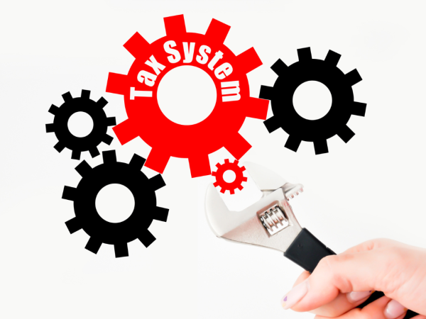 various sized cogs interlinked together, the largest red cog has the words 'TAX SYSTEM' on it, a person is turning the smallest red cog with an adjustable wrench. various sized cogs interlinked together, the largest red cog has the words 'TAX SYSTEM' on it, a person is turning the smallest red cog with an adjustable wrench.
