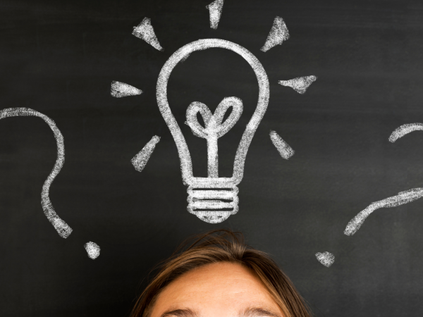 Person stood in front of a chalkboard, the chalkboard has a drawing of a lightbulb and question marks Person stood in front of a chalkboard, the chalkboard has a drawing of a lightbulb and question marks