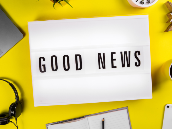 Light up box with the words 'GOOD NEWS' on it, a laptop, headphones, notepad and pen, coffee cup amongst other items are sat on the yellow desk.