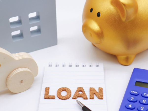 A white desk containing a golden piggy bank, a silver wooden house, a wooden car, the word 'LOAN' and a calculator. 