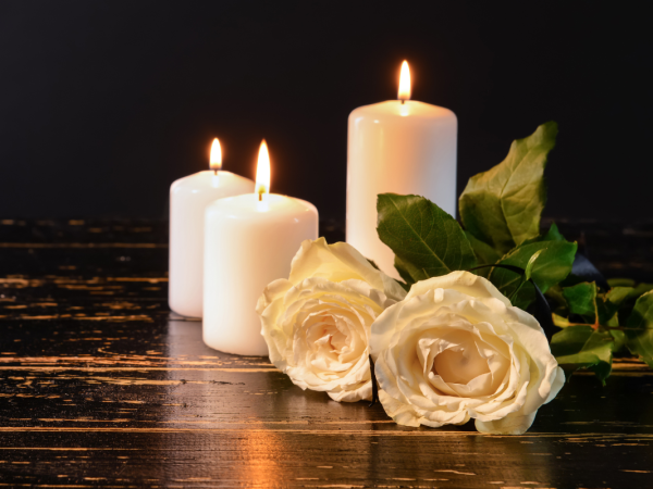 3 white candles lit alongside 2 white roses laid on a dark wooden table. 