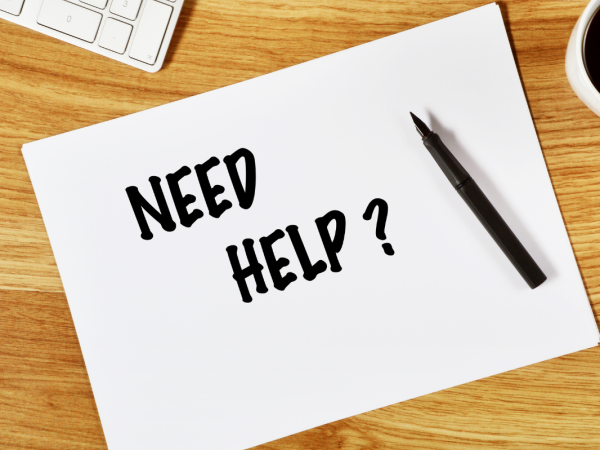 A white sheet of paper on a wooden desk with the words 'NEED HELP' written on it in black ink.