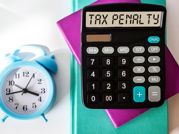 Calculator displaying ‘Tax Penalty’ beside a blue alarm clock and colourful notebooks.