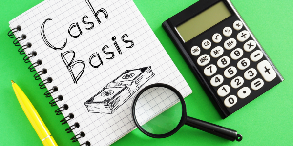 a calculator, a magnifying glass and a pad of squared paper with the words 'Cash basis' written on the top sheet along with an illustration of a stack of money