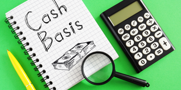 a calculator, a magnifying glass and a pad of squared paper with the words 'Cash basis' written on the top sheet along with an illustration of a stack of money a calculator, a magnifying glass and a pad of squared paper with the words 'Cash basis' written on the top sheet along with an illustration of a stack of money