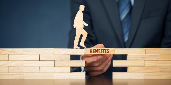 wooden blocks making a bridge with a gap in it, one block is being held in place to allow the wooden figure to cross, on this block the word 'BENEFITS' is written on the side.