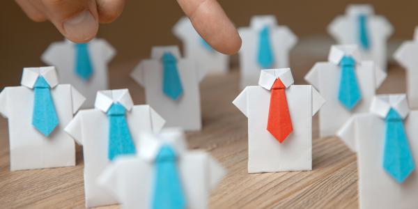 A table of origami shirts made from white paper, each wearing a little blue tie except for one that is wearing a red tie, a person is pointing to the origami shirt wearing the red tie.