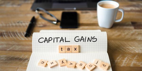 A pad of squared paper with the words 'CAPITAL GAINS' written on it in black ink, wooden scrabble tiles are scattered over the bottom of the paper with the word 'TAX' arranged to spell out 'CAPITAL GAINS TAX' 