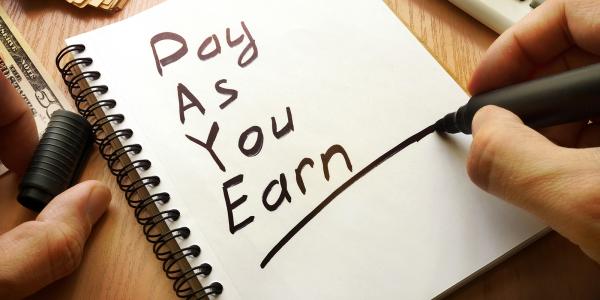 A person writing on a pad of white paper, the words 'PAY AS YOU EARN' are written in black handwriting and underlined.