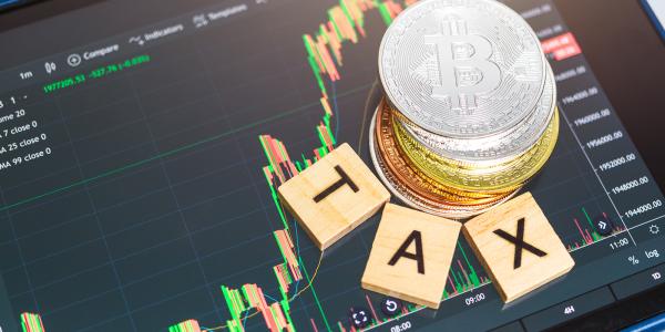 a tablet showing a cryptocurrency graph, a stack of Cryptocurrency, and the word 'TAX' arranged around the stack of coins with wooden scrabble tiles a tablet showing a cryptocurrency graph, a stack of Cryptocurrency, and the word 'TAX' arranged around the stack of coins with wooden scrabble tiles