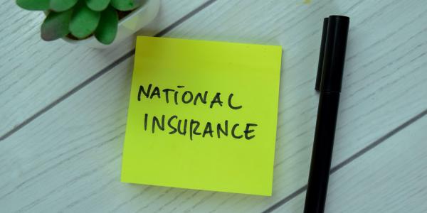 a yellow post it note with the words 'NATIONAL INSURANCE' written on it in black ink, a black pen sits next to this and also a small plant. 