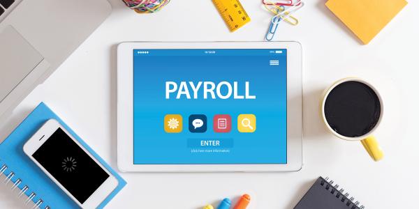 A tablet showing 'PAYROLL' on the screen, the tablet is sat on a desk surrounded with notebooks, a phone, a cup of coffee, paperclips, a ruler, sticky notes and coloured pens. A tablet showing 'PAYROLL' on the screen, the tablet is sat on a desk surrounded with notebooks, a phone, a cup of coffee, paperclips, a ruler, sticky notes and coloured pens.