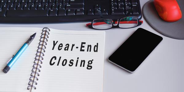 A pad of paper on a desk with the words 'YEAR-END CLOSING' typed on it. also on the deak a pen, a phone, a pair of glasses, a mouse mat and mouse and a keypad can be seen.