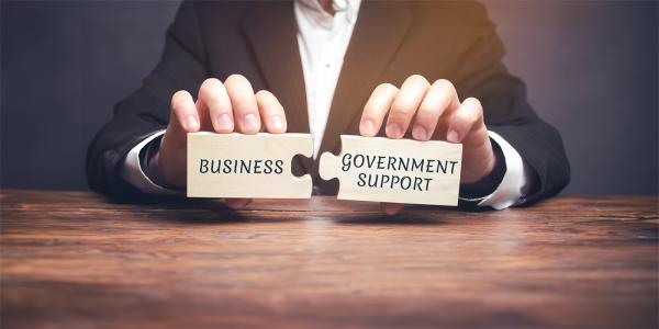 2 pieces of wooden jigsaw being held together by a person, each jigsaw piece has words written on them, one says 'BUSINESS' and the other says 'GOVERNMENT SUPPORT'. 2 pieces of wooden jigsaw being held together by a person, each jigsaw piece has words written on them, one says 'BUSINESS' and the other says 'GOVERNMENT SUPPORT'.
