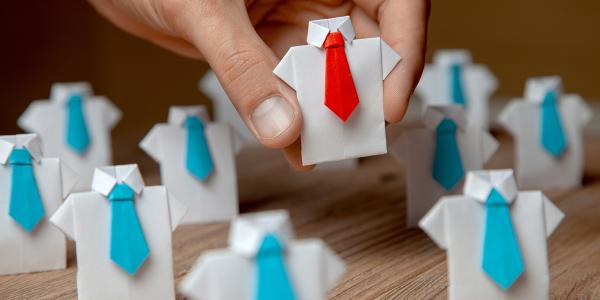 a desk of little white origami shirts wearing coloured tie's, all of which have blue ties except for the one in the persons hand which is wearing a red tie.