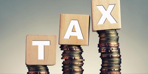 3 piles of coins with a wooden block on top of each pile, each block has a letter on it, together they spell the word 'TAX' 