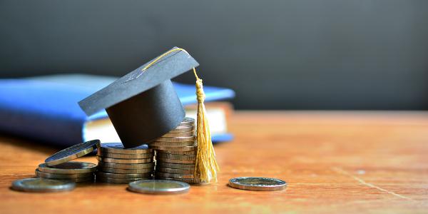 a pile of coins on a wooden table, in the background sits a blue book. On top of the coin pile is a tiny black graduation cap