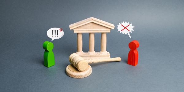 a wooden made figure of a courthouse, a wooden Gavel and 2 small wooden people with speech bubbles above their heads, the green person's speech bubble shows '!!!' the red one's speech bubble is jagged edged and shows 'X'