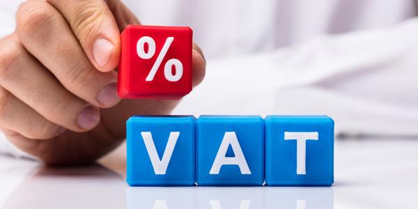 a person stacking blocks, 3 blue blocks sat next to each other read 'VAT' then a further red block on top of these with the '%' sign on it. a person stacking blocks, 3 blue blocks sat next to each other read 'VAT' then a further red block on top of these with the '%' sign on it.