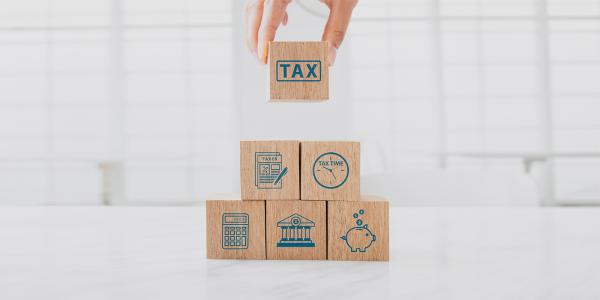 6 wooden building blocks each with a tax related image on such as the word 'TAX' a document of 'TAXES', a clock with the words 'TAX TIME', a calculator, a bank, a piggy bank. 