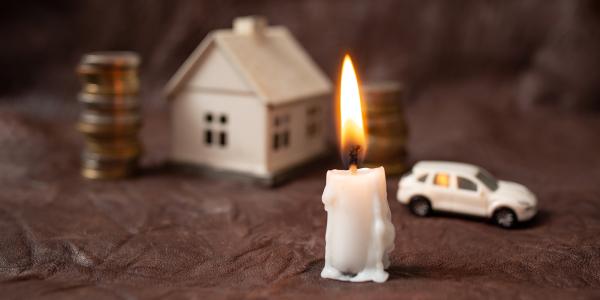 a lit candle, in the background a stack of coins, a house, and a car can be seen. 