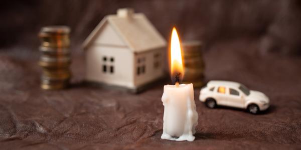 a lit candle, in the background a stack of coins, a house, and a car can be seen. a lit candle, in the background a stack of coins, a house, and a car can be seen.