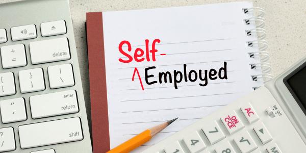 A keyboard, calculator, pencil and a pad of paper with the words 'SELF-EMPLOYED' written on it in black and red ink. 
