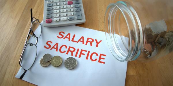 a sheet of white paper with the words 'SALARY SACRIFICE' typed on them in red ink. next to this is a pair of glasses, a calculator an a jar of money tipped on its side, some of the money has made it's way onto the paper. 