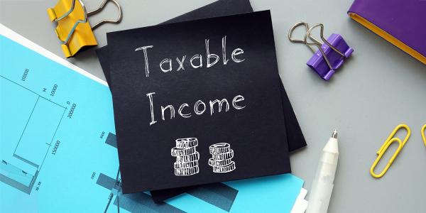 a piece of black paper on a desk surrounded by various stationary and graphs, the paper reads 'TAXABLE INCOME' in white writing along with an image of 2 stacks of coins. 