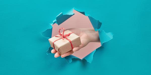 a blue paper wall with a hole in the middle, a persons hand appears through the hole holding out a gift box wrapped in brown paper with red string. 