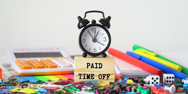 a desk scattered in various stationary, in the centre of the desk is a wooden block with the words 'PAID TIME OFF' and a clock sat on top. a desk scattered in various stationary, in the centre of the desk is a wooden block with the words 'PAID TIME OFF' and a clock sat on top.