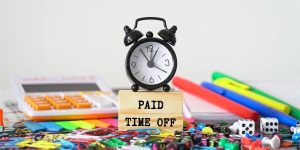 a desk scattered in various stationary, in the centre of the desk is a wooden block with the words 'PAID TIME OFF' and a clock sat on top.