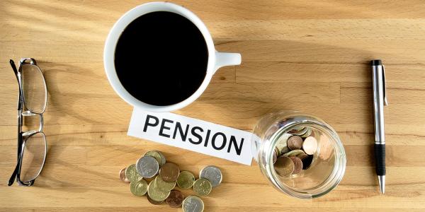 a birds eye view of a table top with a jar of coins, pen, coffee, glasses, loose coins and a piece of paper with the word 'PENSION' in black ink.  