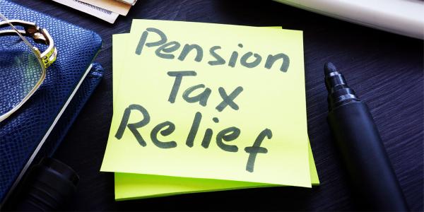 a post-it note against a dark coloured desk that reads 'PENSION TAX RELIEF' next to this is a leather folder, a pair of glasses and a black marker pen. 