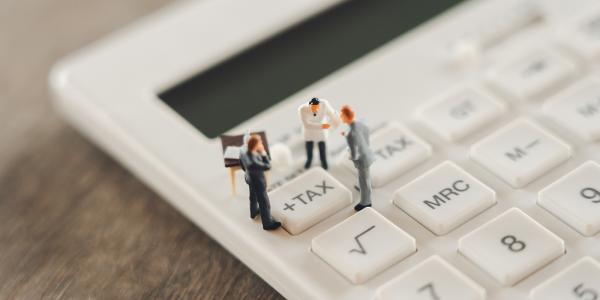 Tiny confused people stood on a calculator, huddled around the '+TAX' button Tiny confused people stood on a calculator, huddled around the '+TAX' button