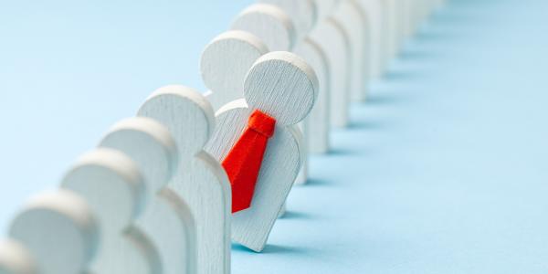 a line of wooden people, one wearing a little red tie is poking out from the line. 