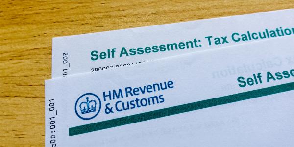 2 sheets of paper on a wooden table, laid on top of each other but slightly overlapping, these are letters from HMRC the front letter shows the HMRC logo in the top left corner, the one behind shows the words 'SELF ASSESSMENT: TAX CALCULATION'