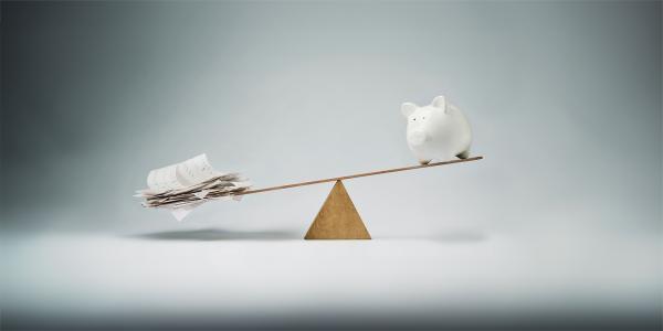 a man made see saw against a neutural coloured background. on one side is a piggy bank, the other side has papers. Image shows the papers are heavier that the piggy bank. 