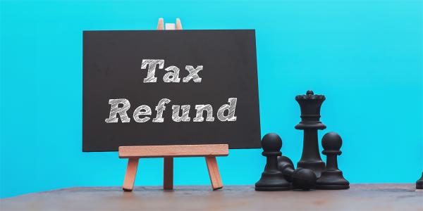 A wooden easel with a chalk board propped on it, the words 'TAX REFUND' are written in white chalk. next to this are some black chess pieces. Behind is a light blue background.