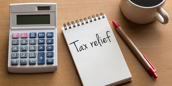 a desk with a calculator, a pen and a cup of coffee, in the centre is a notepad with the words 'TAX RELIEF' written on it. 