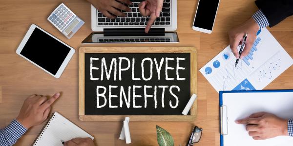 3 people sat around a desk, on the desk is various paperwork, stationary and electronic devices, in the centre of the desk is a chalkboard with the words 'EMPLOYEE BENEFITS' written on it in white chalk. 