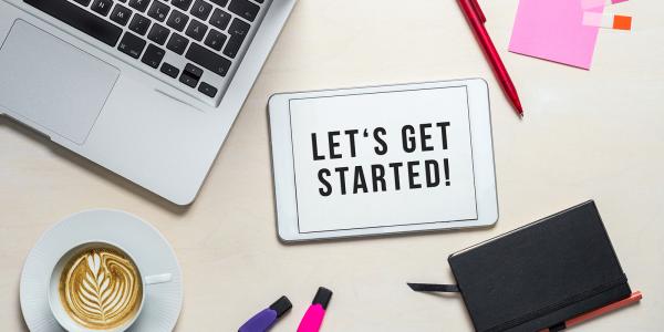 a desk with a laptop, pen, coffee, diary, highlighters and a tablet with the words 'LETS GET STARTED'  on the screen. 