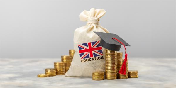 A white sack with a union jack printed on the front, the word 'EDUCATION' typed underneath in back text. next to the sack are stacks of coins, one stack is wearing a tiny graduation cap. 