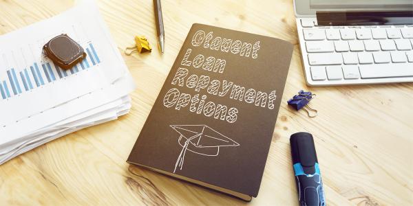 A desk with a keyboard, paperwork, various stationary and a black book with a graduation cap drawn on the front in white pen, the text on the front of the book reads 'STUDENT LOAN REPAYMENT OPTIONS'. 