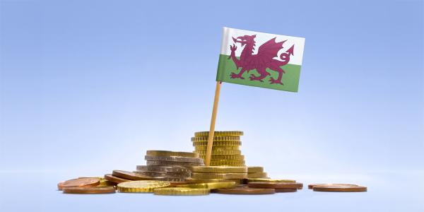 a pile of coins against a blue background, a Welsh flag is stood amongst the pile of coins. 