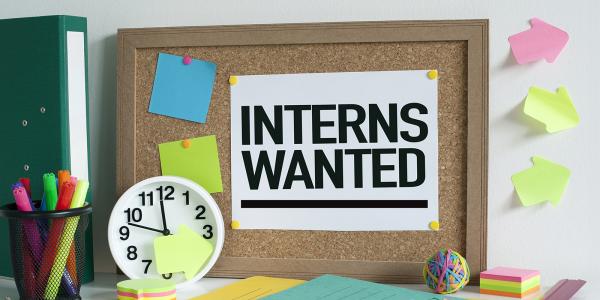 a message board with the words 'INTERNS WANTED' pinned to it,  across the desk various stationary can be seen and a clock. 
