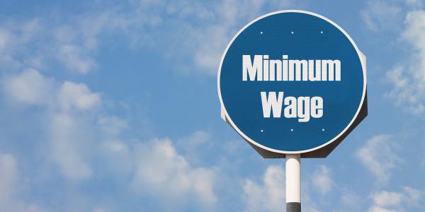 a blue sky with clouds and a blue circular road sign with the words 'MINIMUM WAGE' written on it in white text. 