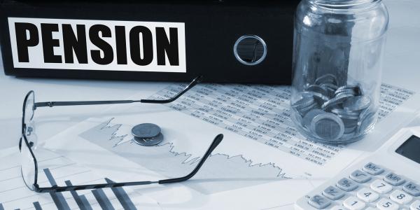 a thick black folder with the word 'PENSION' written on the spine sat on a desk with a pair of glasses, paperwork, a calculator and a jar of coins.